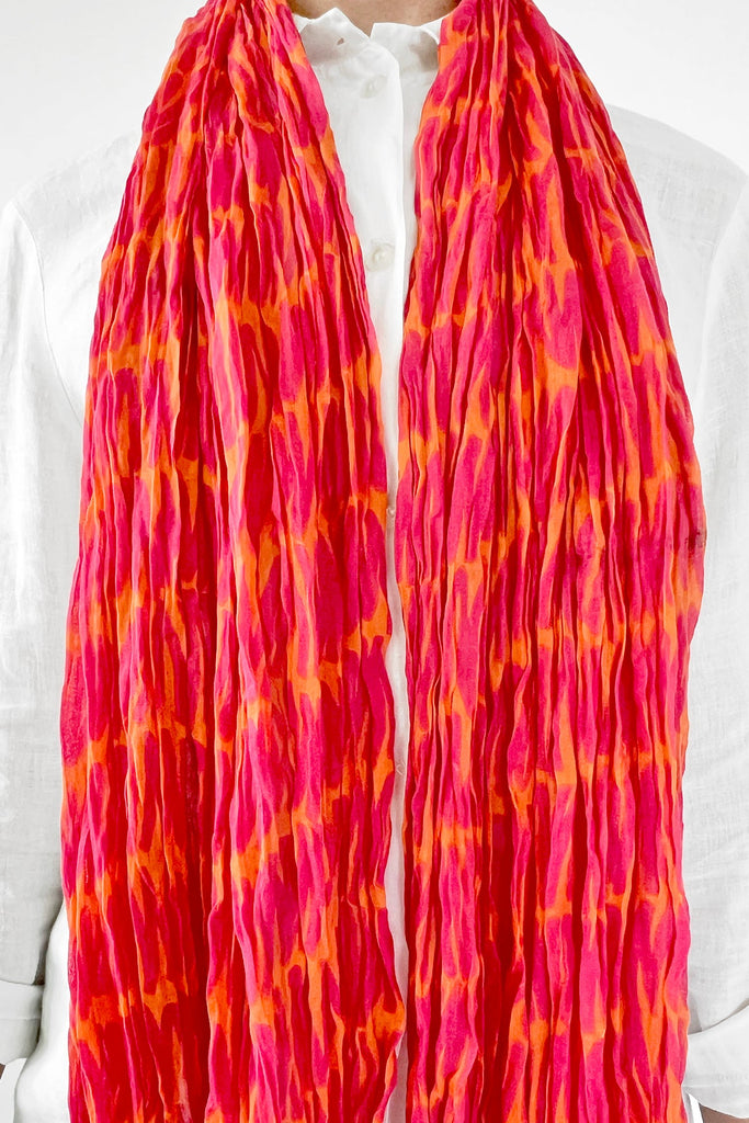 A person wearing a white shirt is shown with the See Design Cotton Scarf, featuring long, crinkled cotton in vibrant red and orange, draped around their neck.