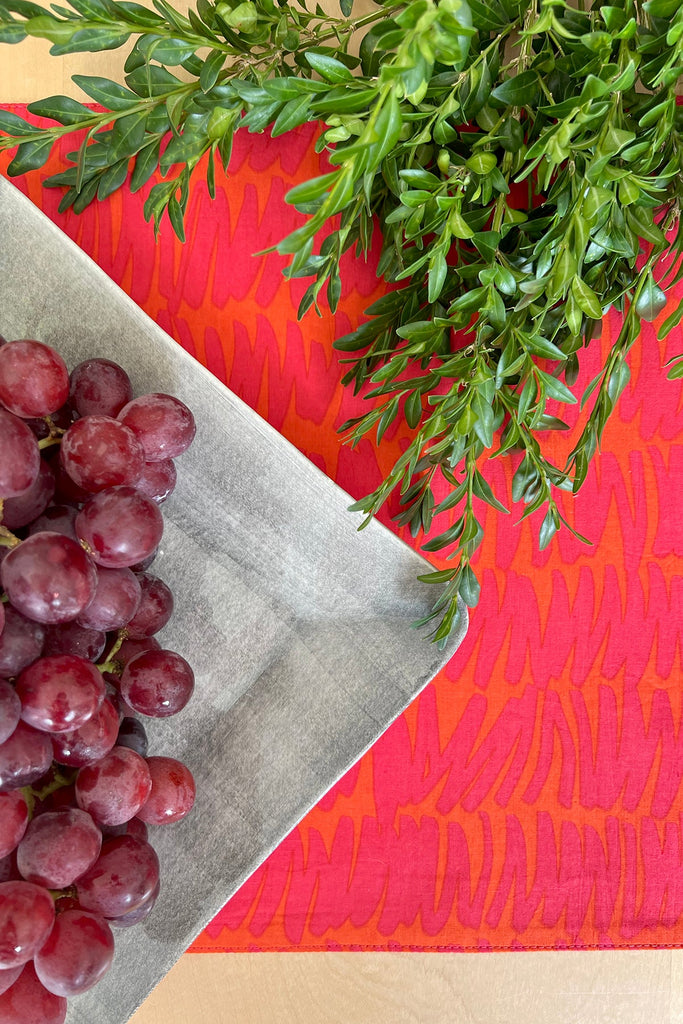 A bunch of red grapes on a gray plate next to green leafy stems, set on a red patterned surface, enhances the dining experience when paired with See Design placemats and matching napkins.