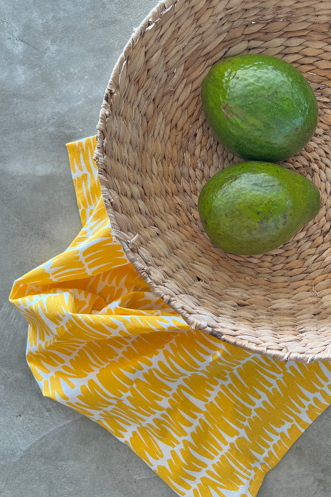 A wicker basket with two avocados and a See Design Tea Towels (Set of 2).