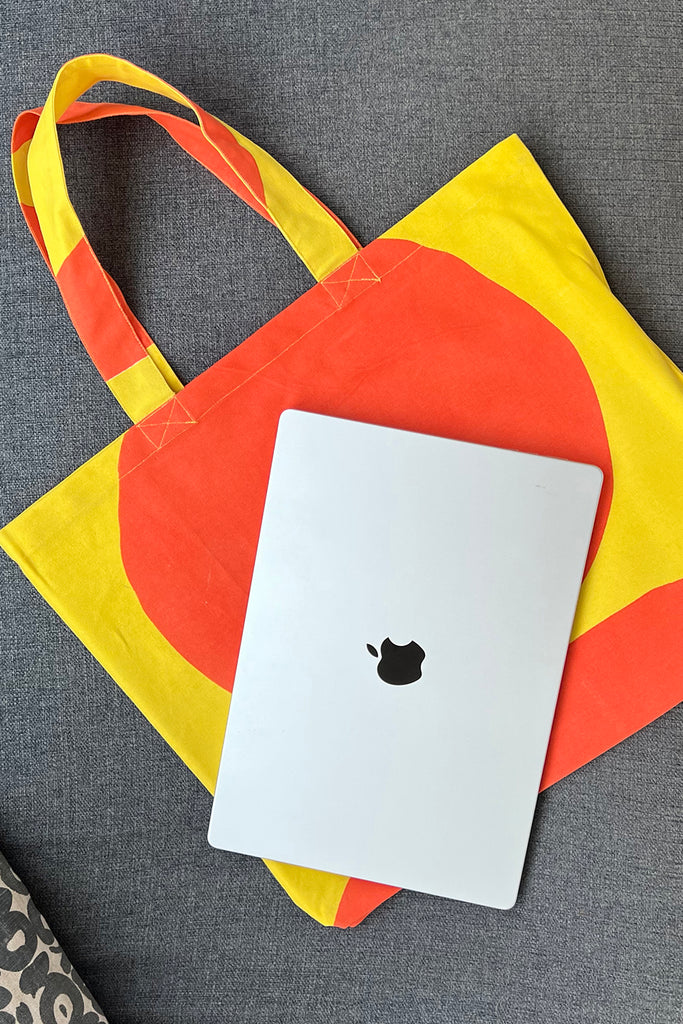 A laptop with an Apple logo rests on a See Design Graphic Tote Bag featuring multicolored cotton canvas with yellow and orange patterns, all placed on a light gray surface.