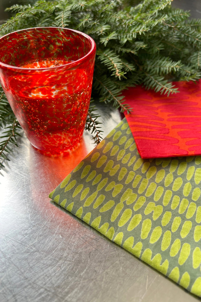 A red speckled glass sits on a table adorned with lush green foliage and two pieces of fabric featuring bold patterns—one is green with yellow ovals and the other is red with orange lines. The vibrant setting is completed by See Design's Cocktail Napkins (Set of 4), adding a burst of color to the scene.