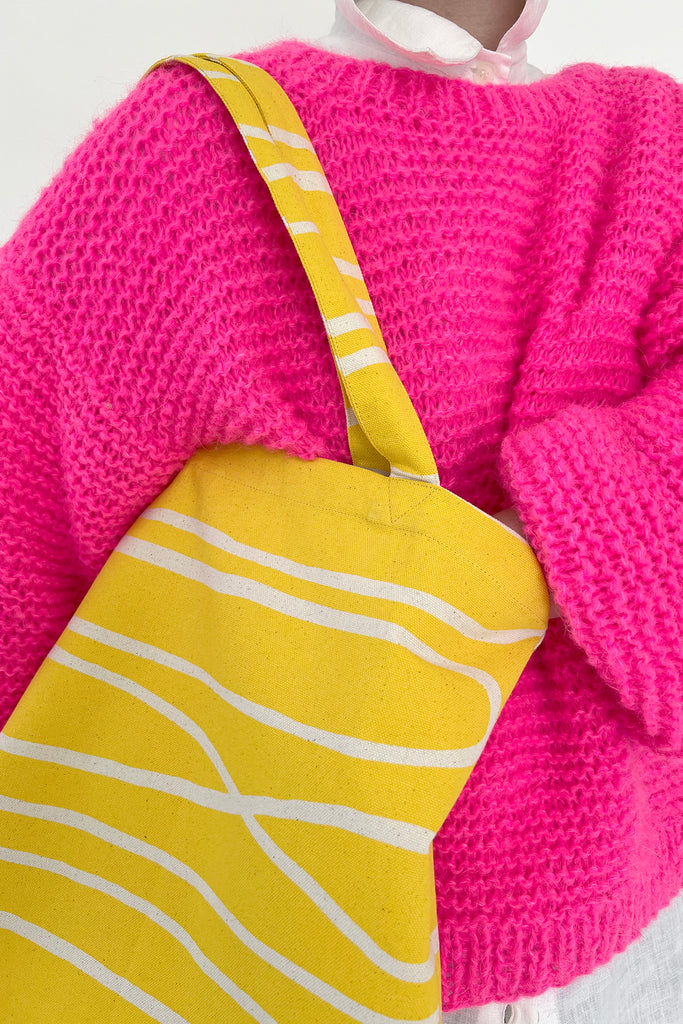 Person wearing a bright pink knit sweater and holding the Easy Tote Bag by See Design, a lightweight cotton canvas tote bag adorned with white abstract lines.