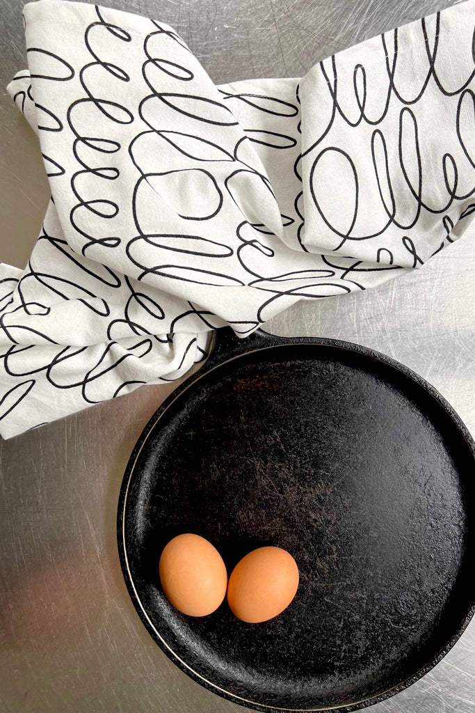 A black frying pan with two eggs on it, perfect for cleaning and drying See Design Tea Towels (Set of 2).