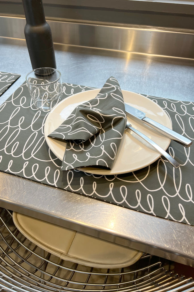 A plate, glass, fork, and knife are arranged on See Design Placemats (Set of 2) with a matching napkin, set atop a metal countertop above a wire shelf and stools for an inviting dining experience.