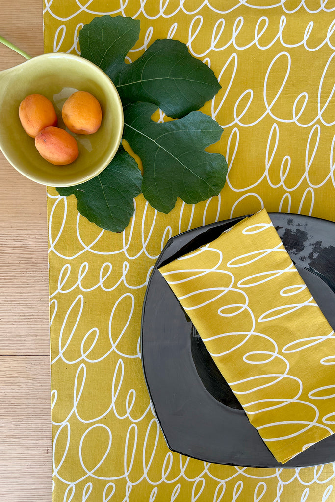 A yellow See Design table runner on a table with a bowl of fruit on it, showcasing vibrant prints and colors.