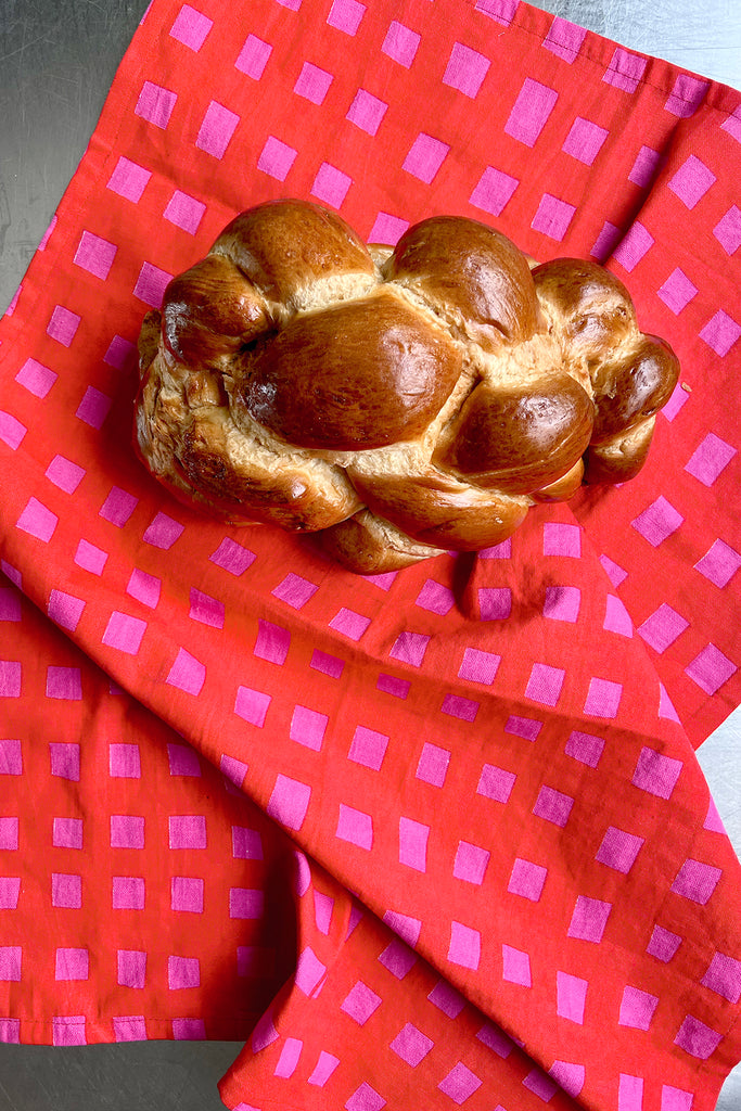 A braided loaf of challah bread rests on a See Design Tea Towel (Set of 2) featuring a red and pink pattern, placed atop a metallic surface.