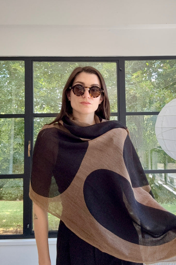 A woman wearing sunglasses and a brown and black See Design Wool Scarf stands indoors in front of large windows with greenery visible outside.