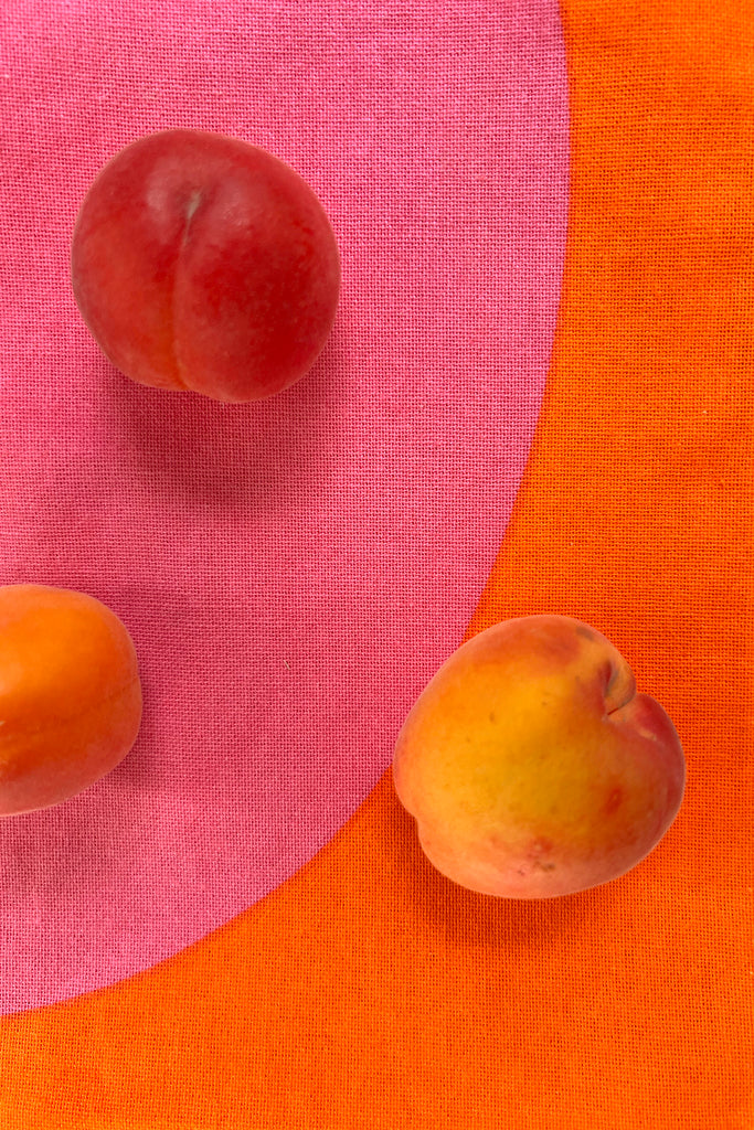 Three ripe peaches and apricots on a fabric background with intersecting pink and orange sections, reminiscent of hand painted designs perfect for See Design Tea Towels (Set of 2) in a welcoming kitchen setting.