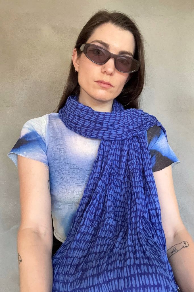 A person wearing sunglasses, a See Design Cotton Scarf with blue patterns, and a short-sleeve shirt stands against a plain, light-colored background.