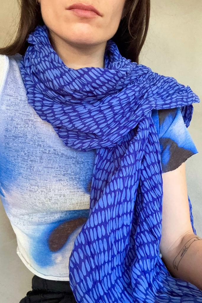 A person wearing a blue and white patterned top and a See Design Cotton Scarf with blue oval spots; a tattoo is visible on their forearm.