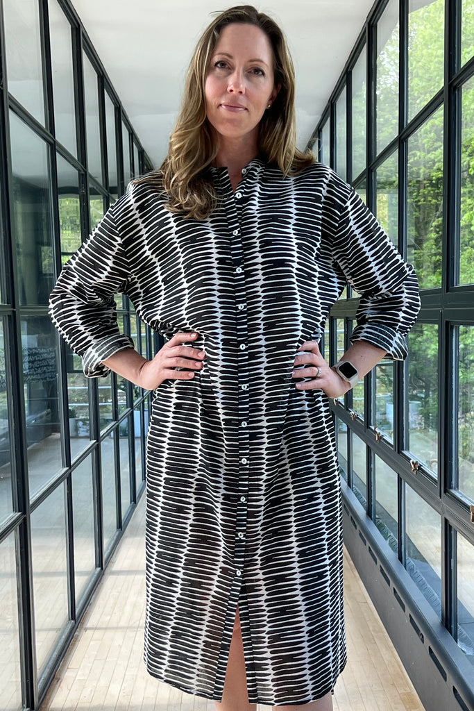 A woman in a See Design striped shirt dress standing confidently in a glass-walled corridor.