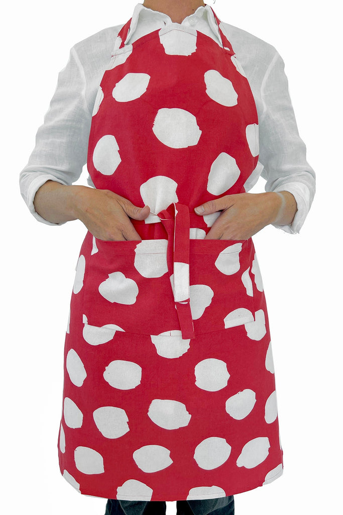A person wears a red See Design Apron with large white polka dots over a white long-sleeve shirt, hands in the front pocket—perfect for fans of hand-painted style.