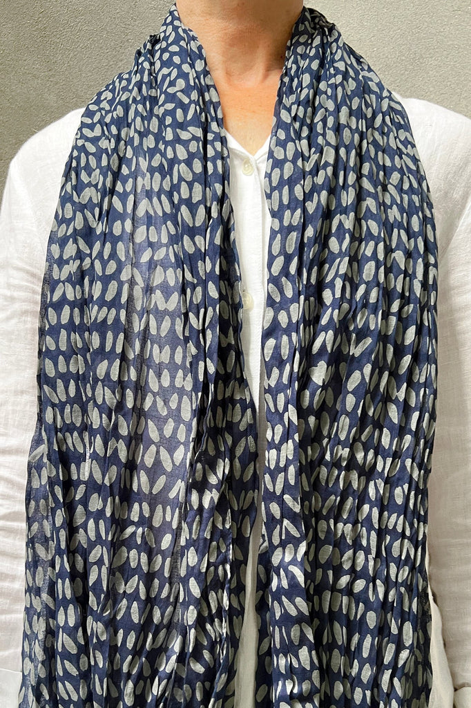 Person wearing a white shirt and a See Design Cotton Scarf in blue crinkled cotton featuring a white oval pattern. The background is plain gray.