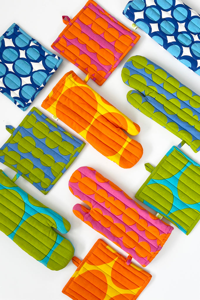 See Design's Oven Mitt & Potholder Set, crafted from soft cotton, showcases an elegant combination of abstract circular patterns in blue, orange, green, and pink on a white background.