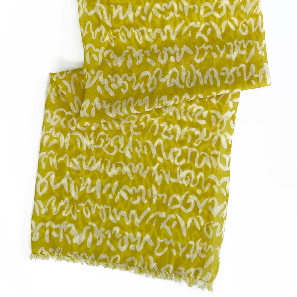 A yellow Wool Scarf by See Design, featuring white abstract squiggly lines and lightly frayed edges, is displayed on a white surface.