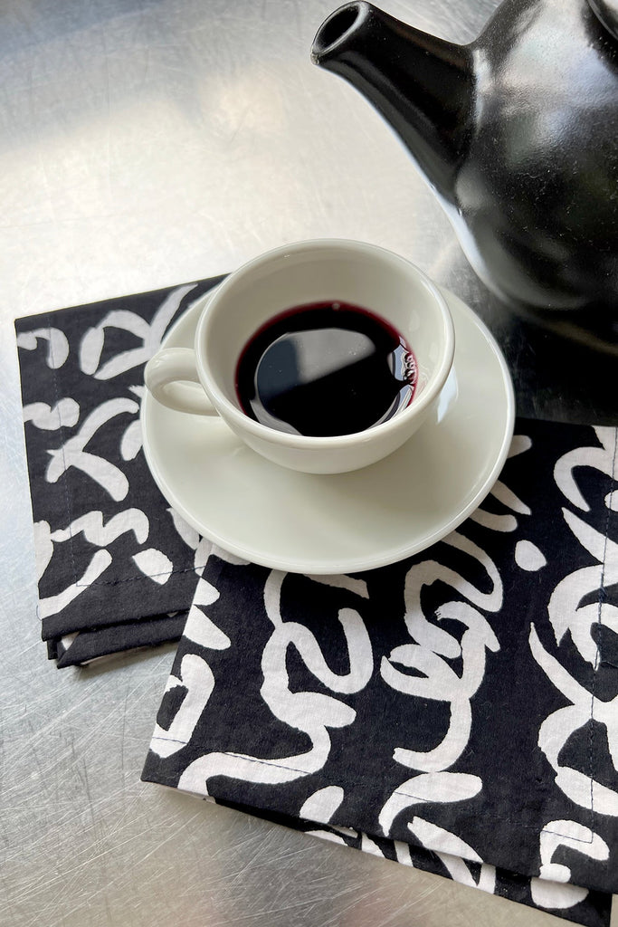 A white teacup filled with dark liquid sits on a white saucer atop See Design's black and white patterned cocktail napkins, next to a black teapot.