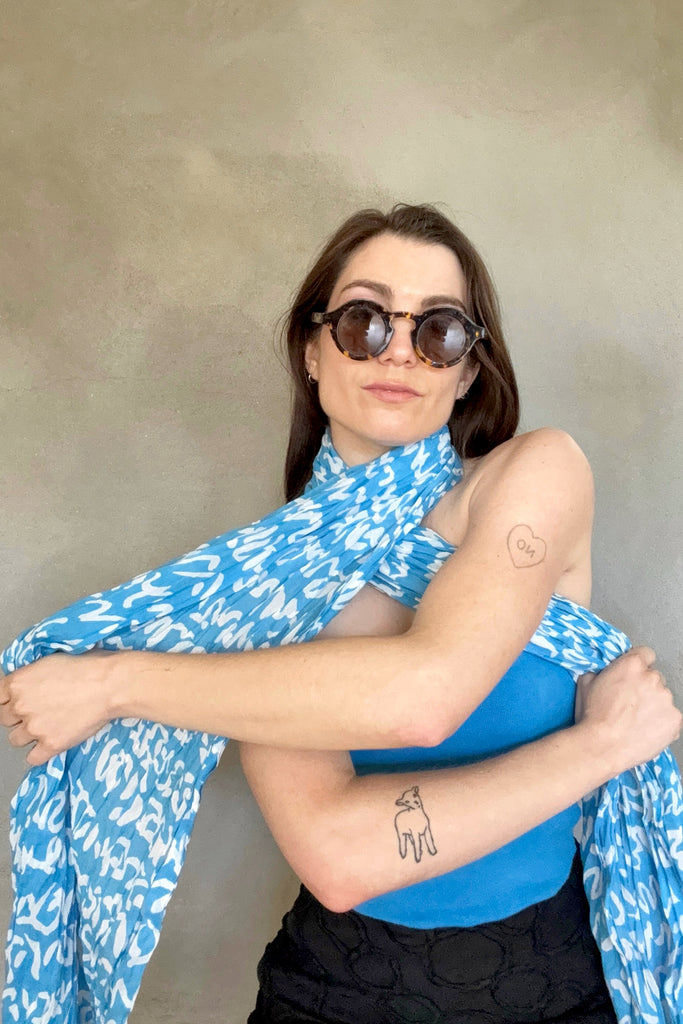 A woman wearing See Design's blue patterned Cotton Scarf, round sunglasses, and a blue top, with tattoos on both arms, stands against a neutral background.