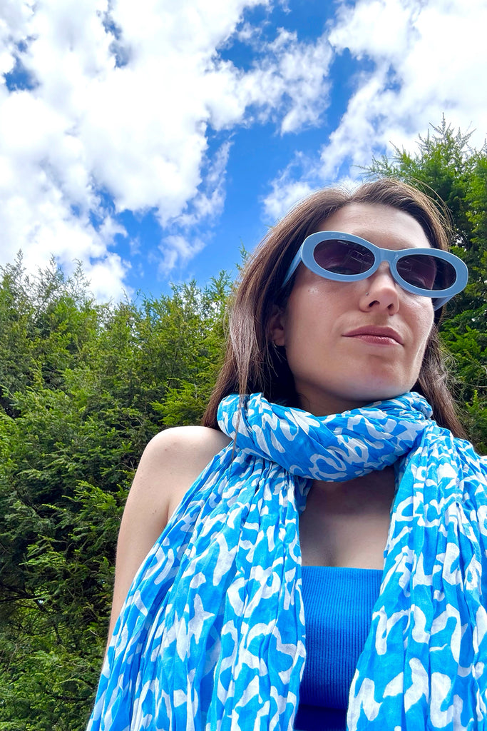 A woman stands outdoors in front of green trees and a partly cloudy sky, wearing blue sunglasses, a blue top, and a See Design Cotton Scarf.