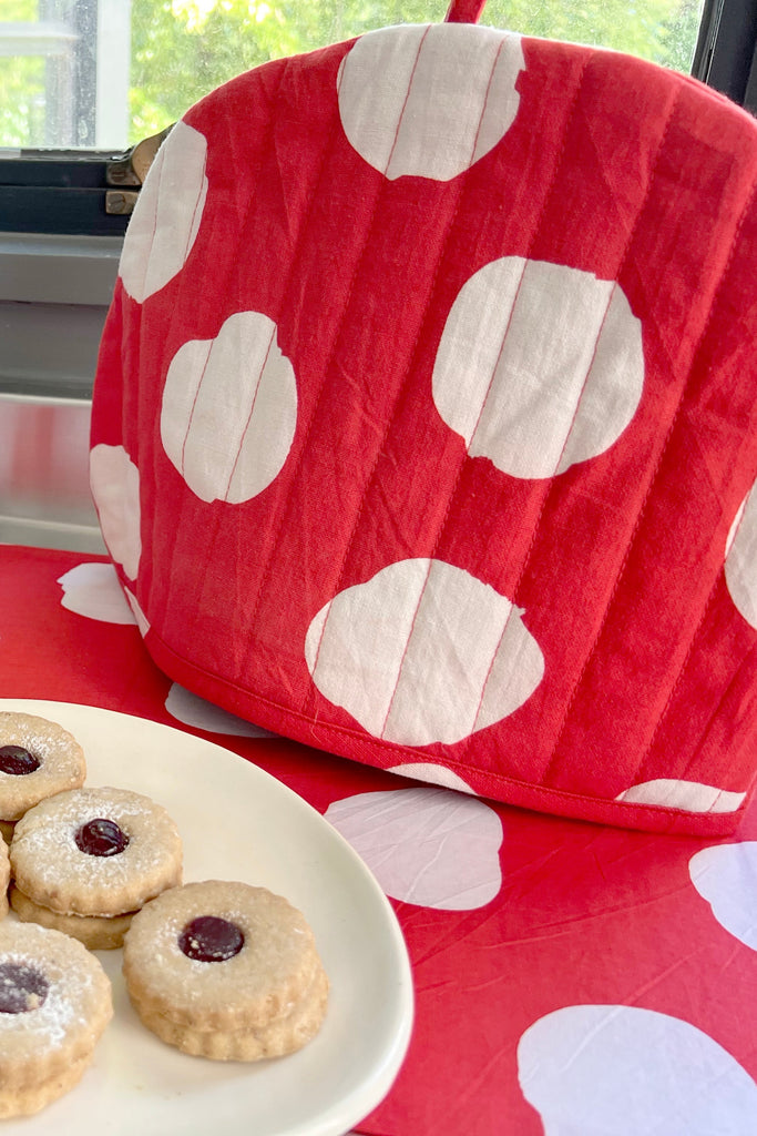 A See Design Tea Cosy with large white spots sits beside a plate of round jam-filled cookies on a red polka dot tablecloth, making it perfect for your fall home collection.