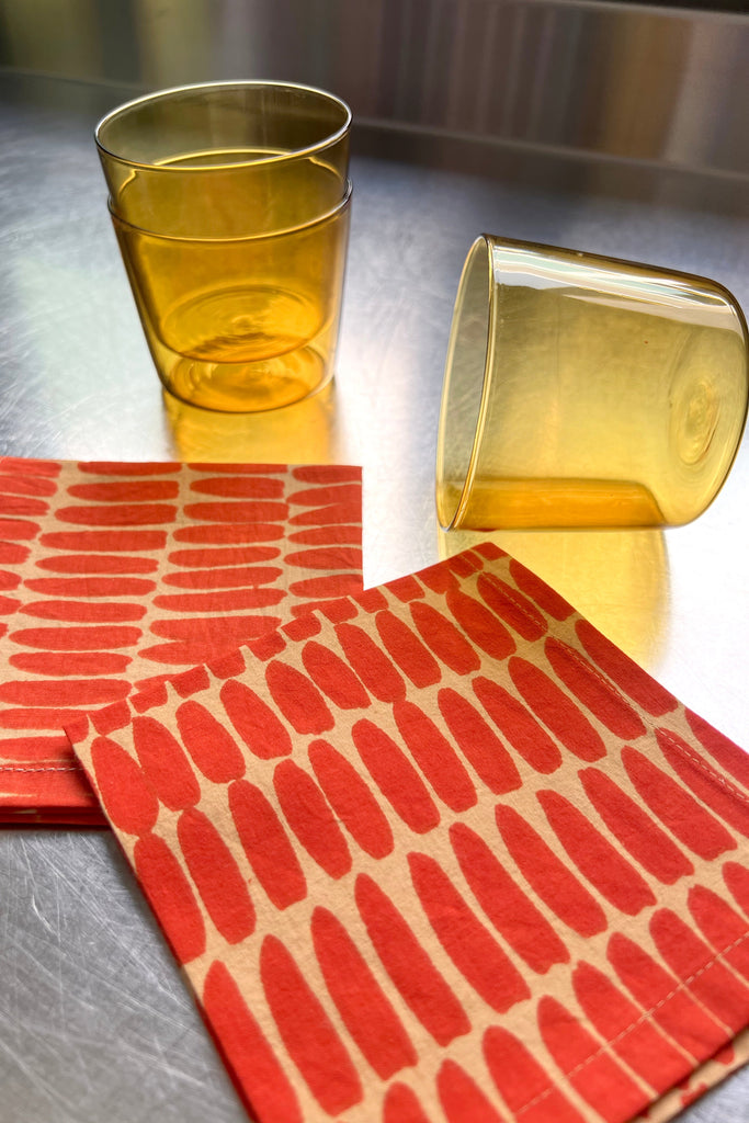 Two stacked amber-colored glasses, one tipped over, sit beside a pile of Cocktail Napkins (Set of 4) by See Design on a stainless steel surface.
