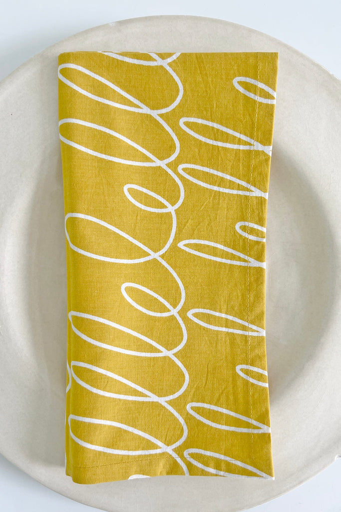 A hand-painted yellow and white cotton napkin (Set of 4) by See Design on a white plate.