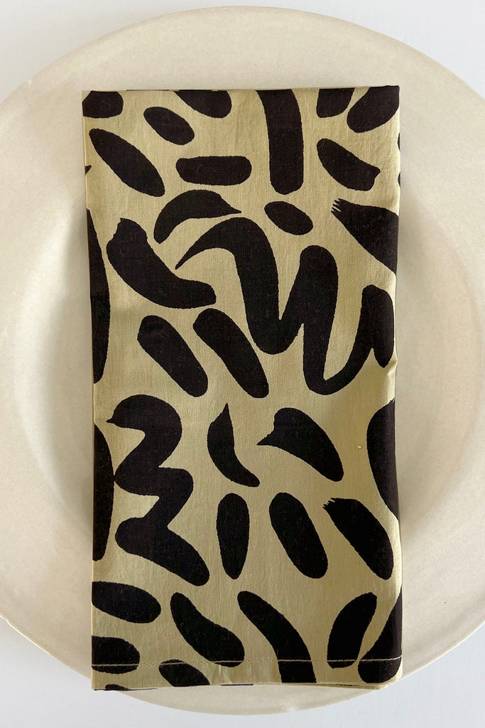 A vibrant black and gold cotton napkin with hand-painted designs on a plate replaced with "See Design Napkins (Set of 4)".