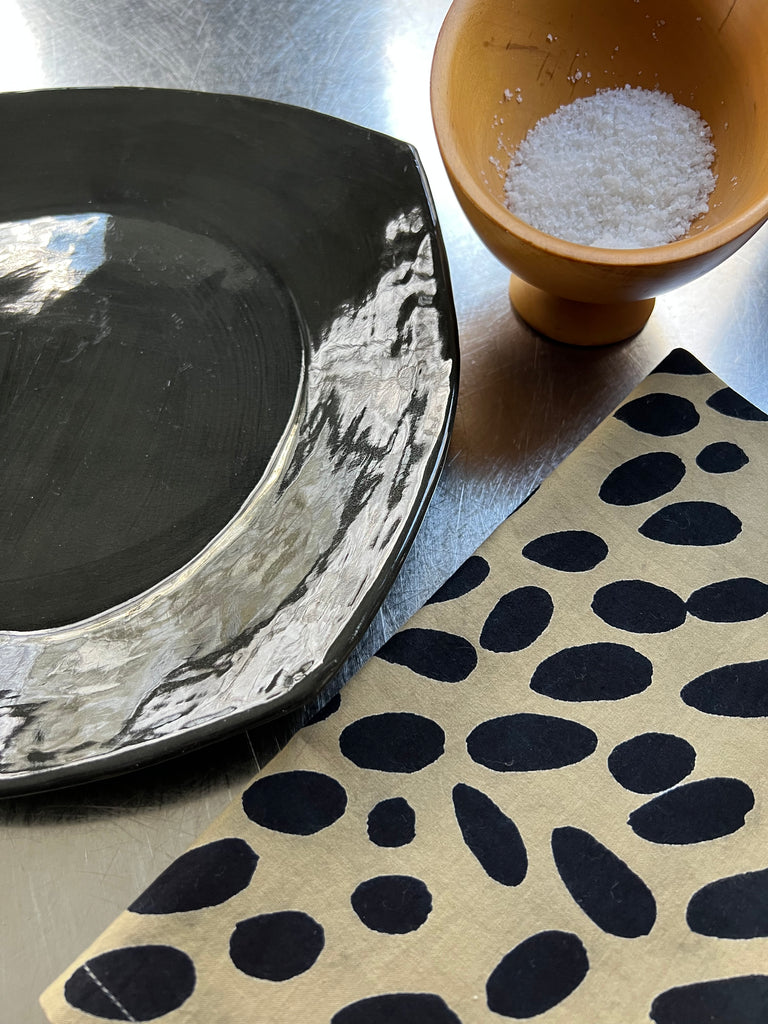 A black and white plate with a bowl of salt next to it, set on See Design colorful table linens.